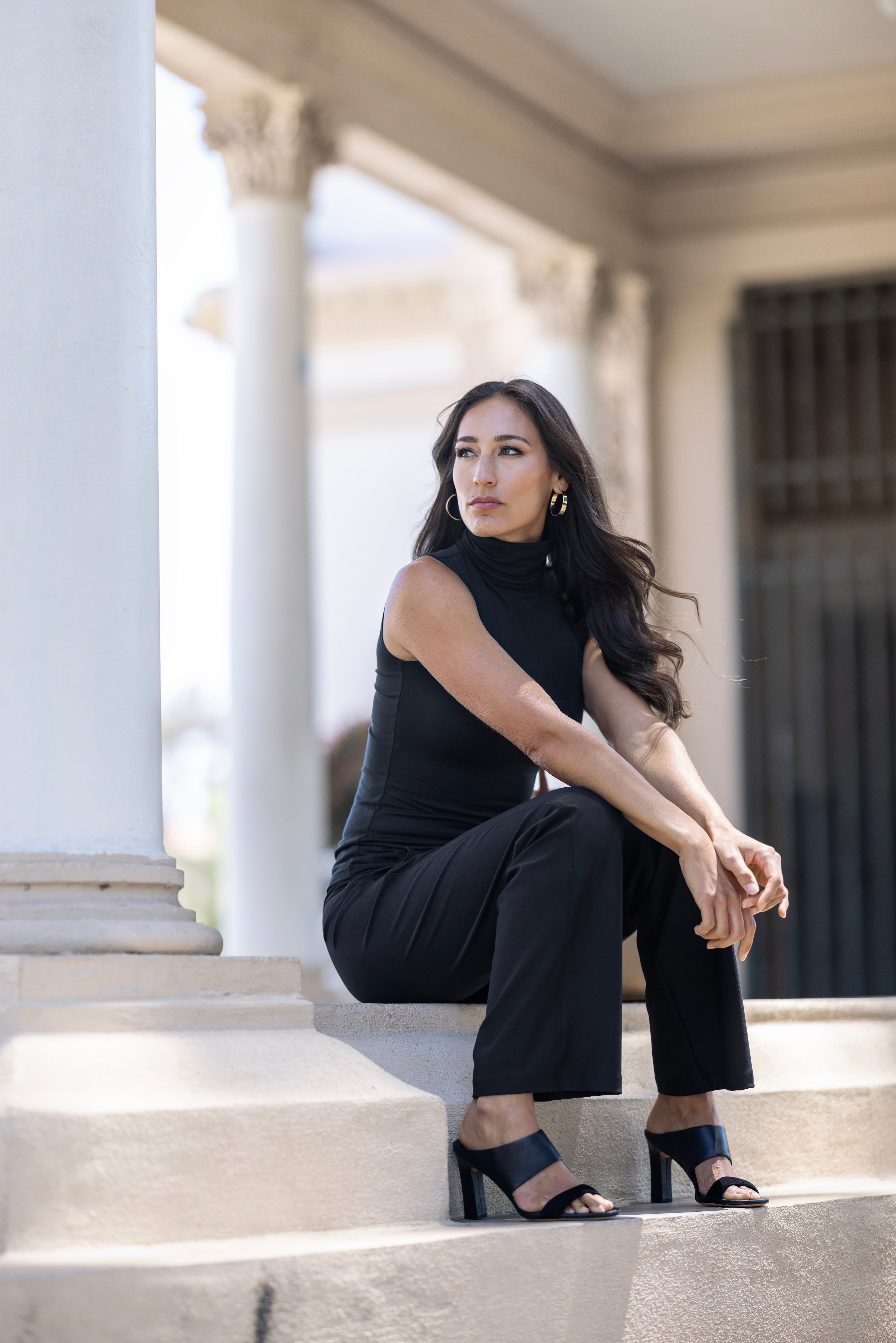 Model sitting wearing a sleeveless black top & black pants.  Beautifully accessories with sandals & earrings for a relaxing day.