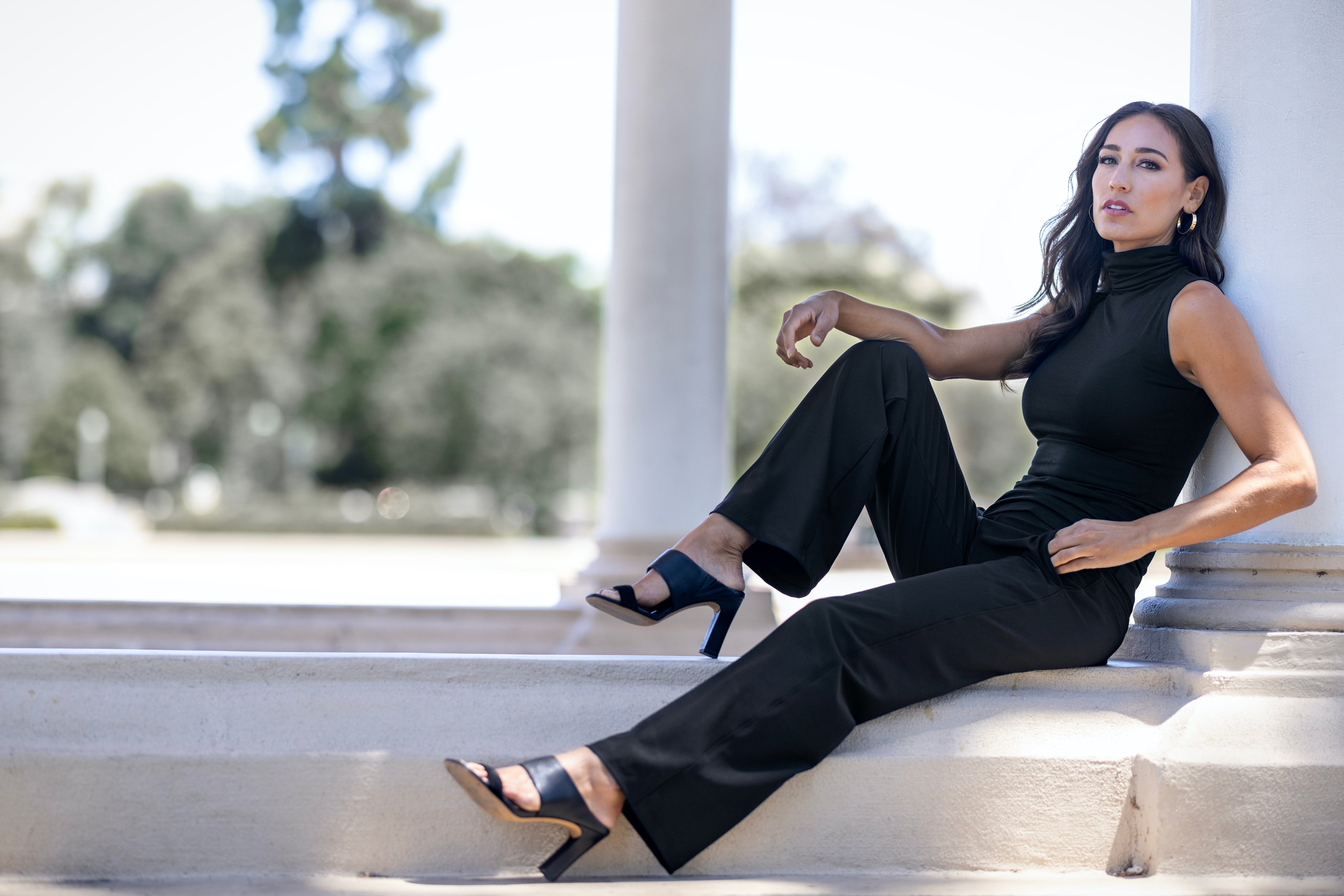 Model sitting wearing a sleeveless black top & black pants.  Beautifully accessories with sandals & earrings for a relaxing day.